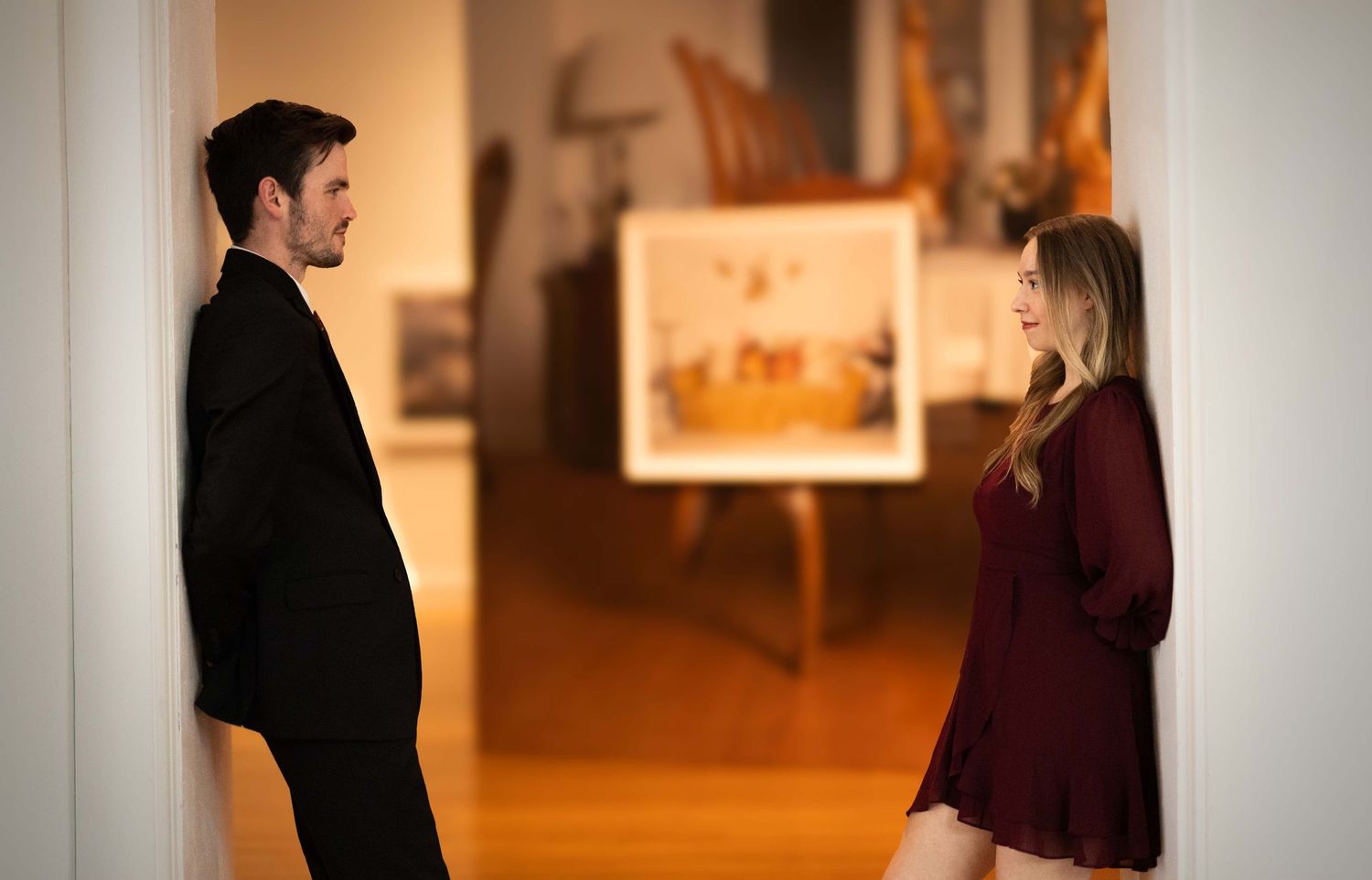 A man and woman in formal attire face each other in a softly lit art gallery hallway, sharing a quiet moment.