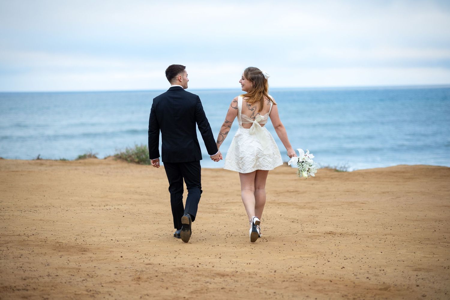 Newlywed couple holding hands walking along sandy coastal bluff with ocean backdrop, bride in short white dress holding bouquet.