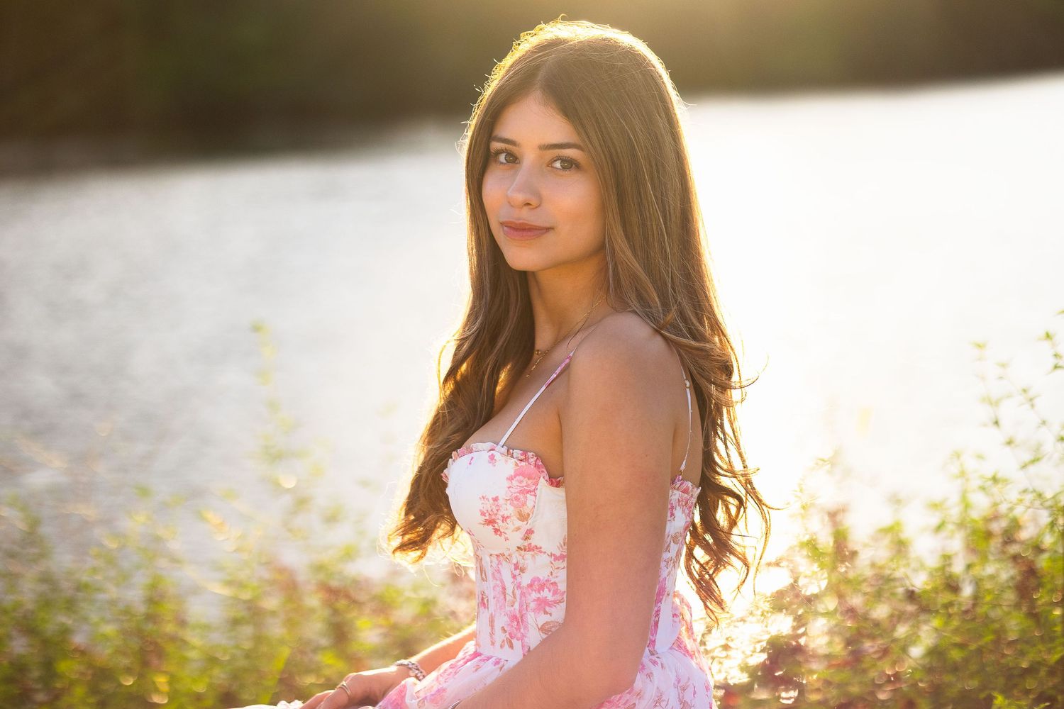 Beautiful young woman in a pink floral sundress standing near a golden sunset river.