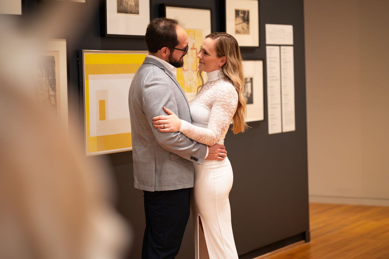 A romantic couple embraces and nearly kisses in front of artwork displayed on a gallery wall.