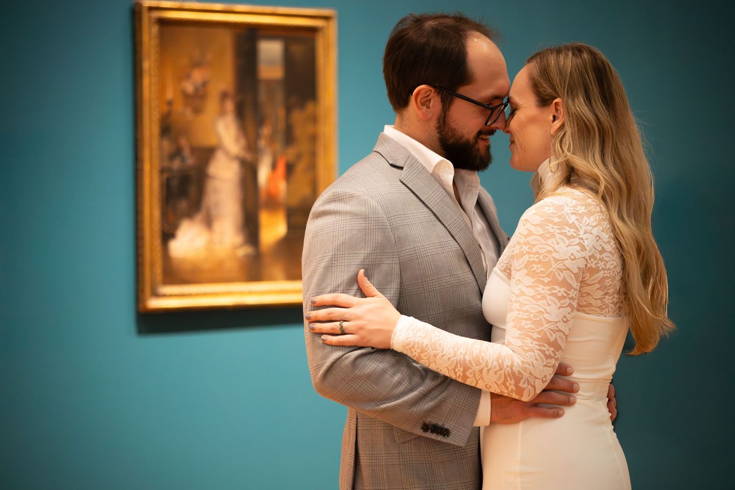Couple in wedding attire embracing romantically in front of a classic painting in a teal-walled art museum gallery.