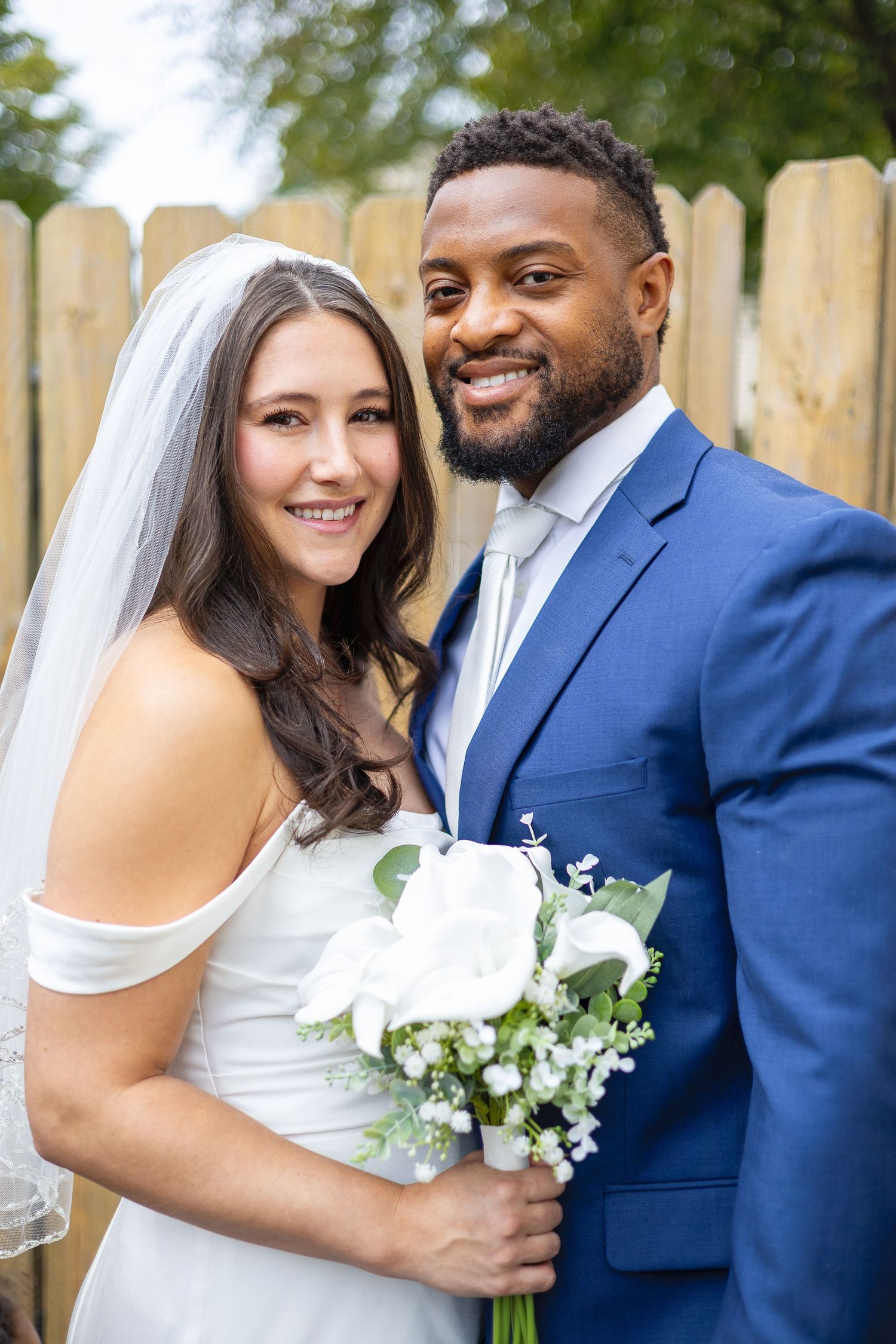 Smiling interracial newlywed couple posing outdoors, bride in white gown holding bouquet.