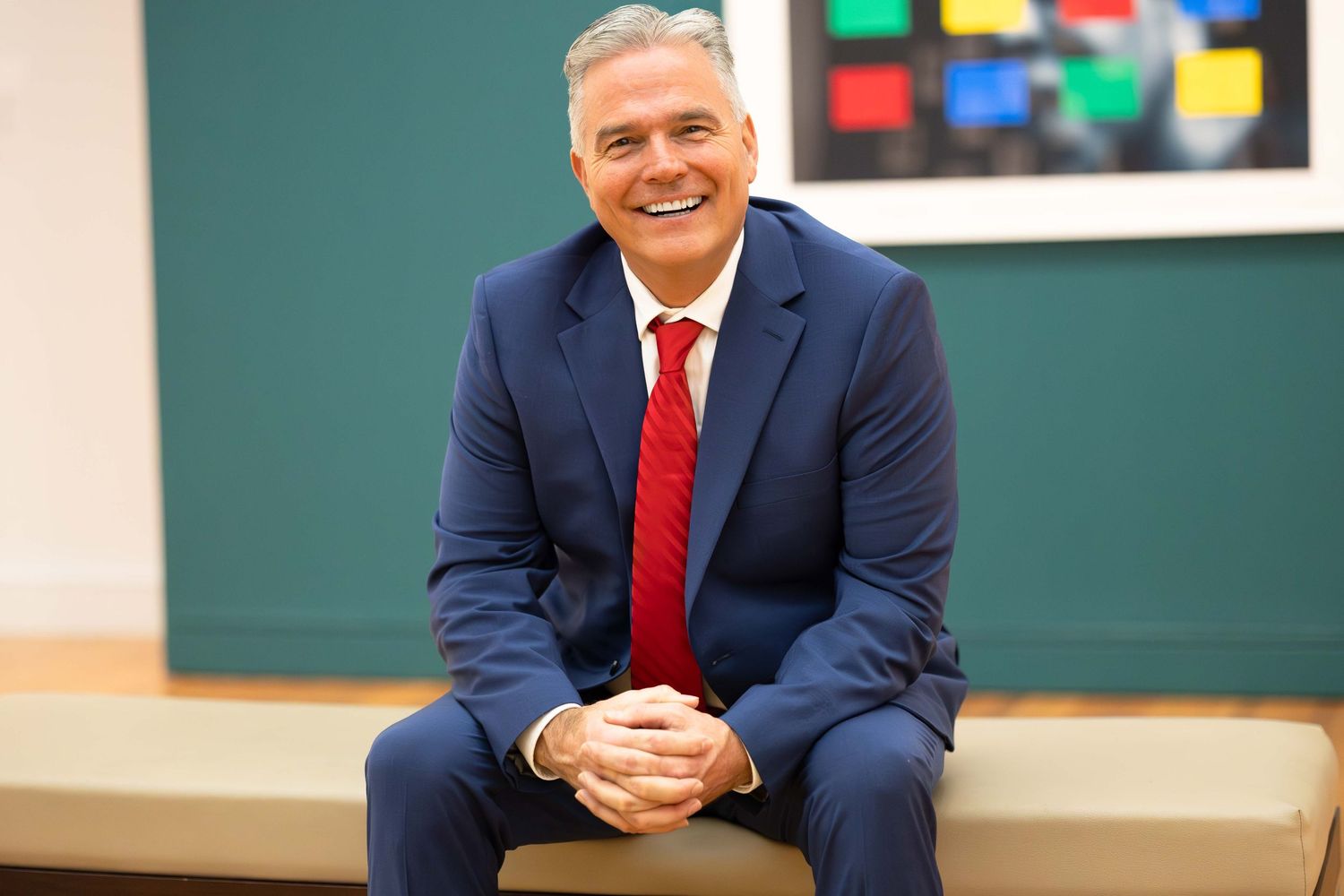 Professional man in navy blue suit and red tie sitting and smiling in modern office setting.