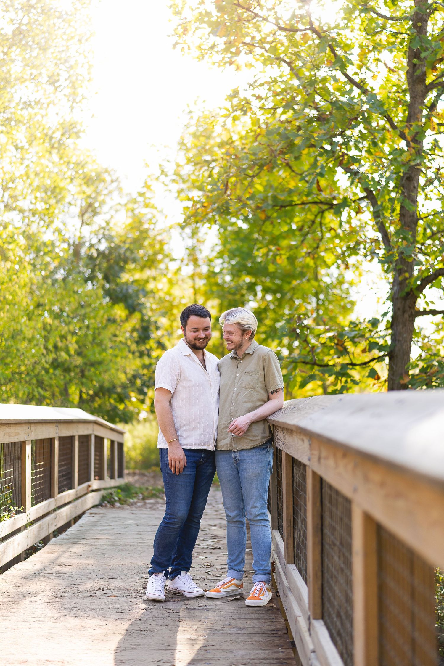 A happy same-sex couple stands close together smiling on a sunny wooden bridge surrounded by trees.