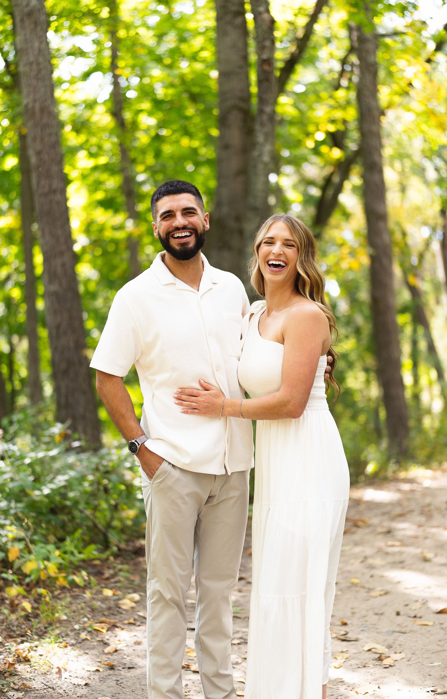 A smiling couple in coordinating white and beige outfits poses together on a wooded path surrounded by lush green trees.