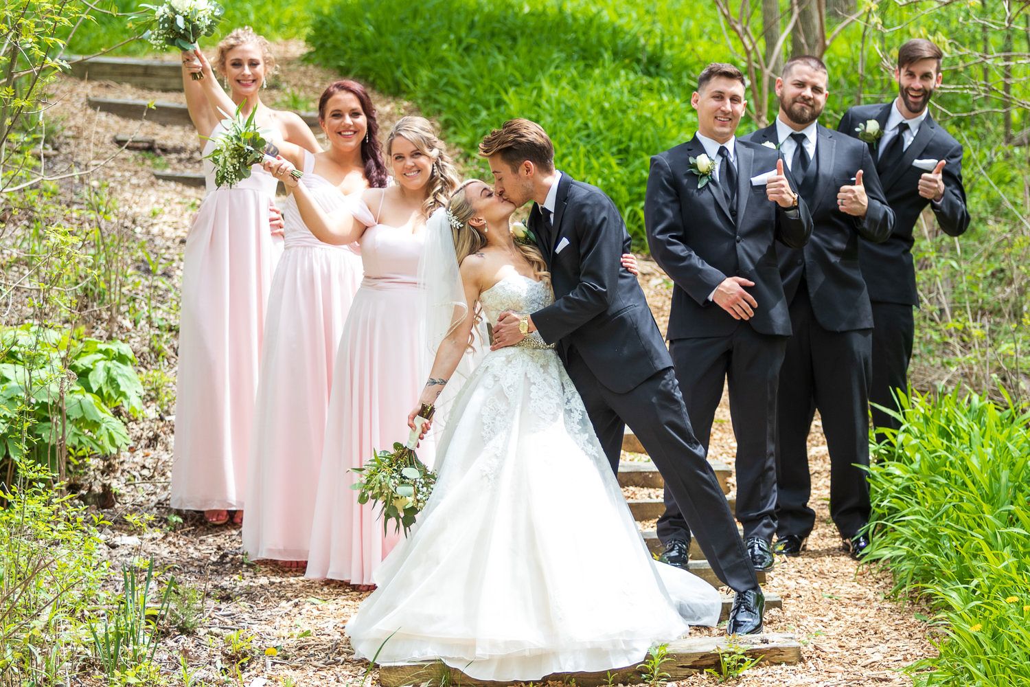 Wedding party poses on a garden path, bride and groom kissing while bridesmaids in pink and groomsmen in black suits celebrate.