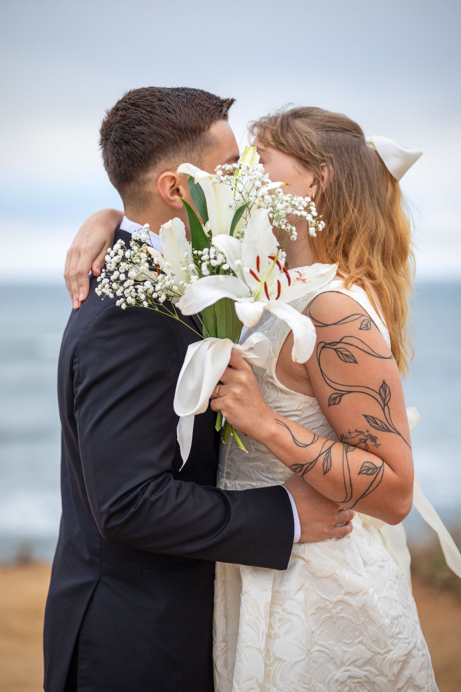 A newlywed couple nearly kisses while holding a white lily bouquet on a beach with ocean views.