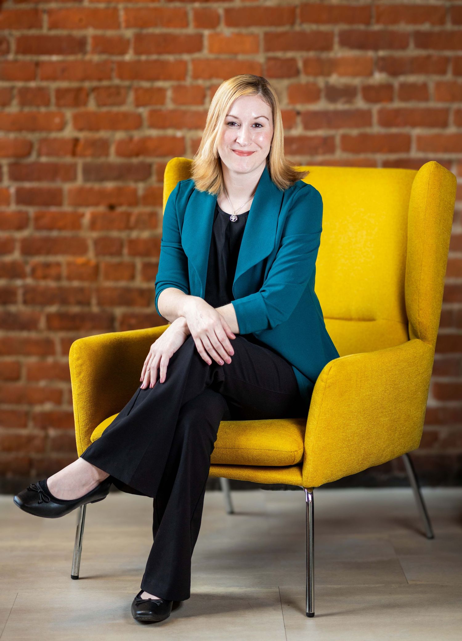 Professional woman in teal blazer sitting in yellow chair against brick wall.