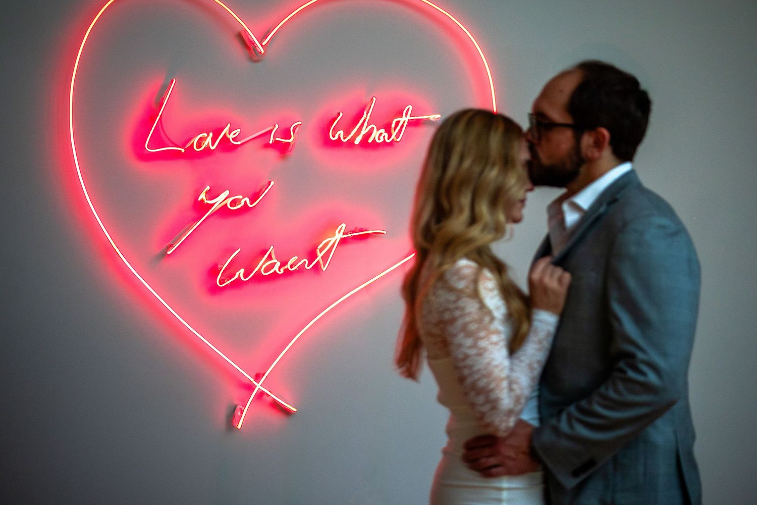 Couple kissing in front of a pink neon heart sign reading Love is What You Want.