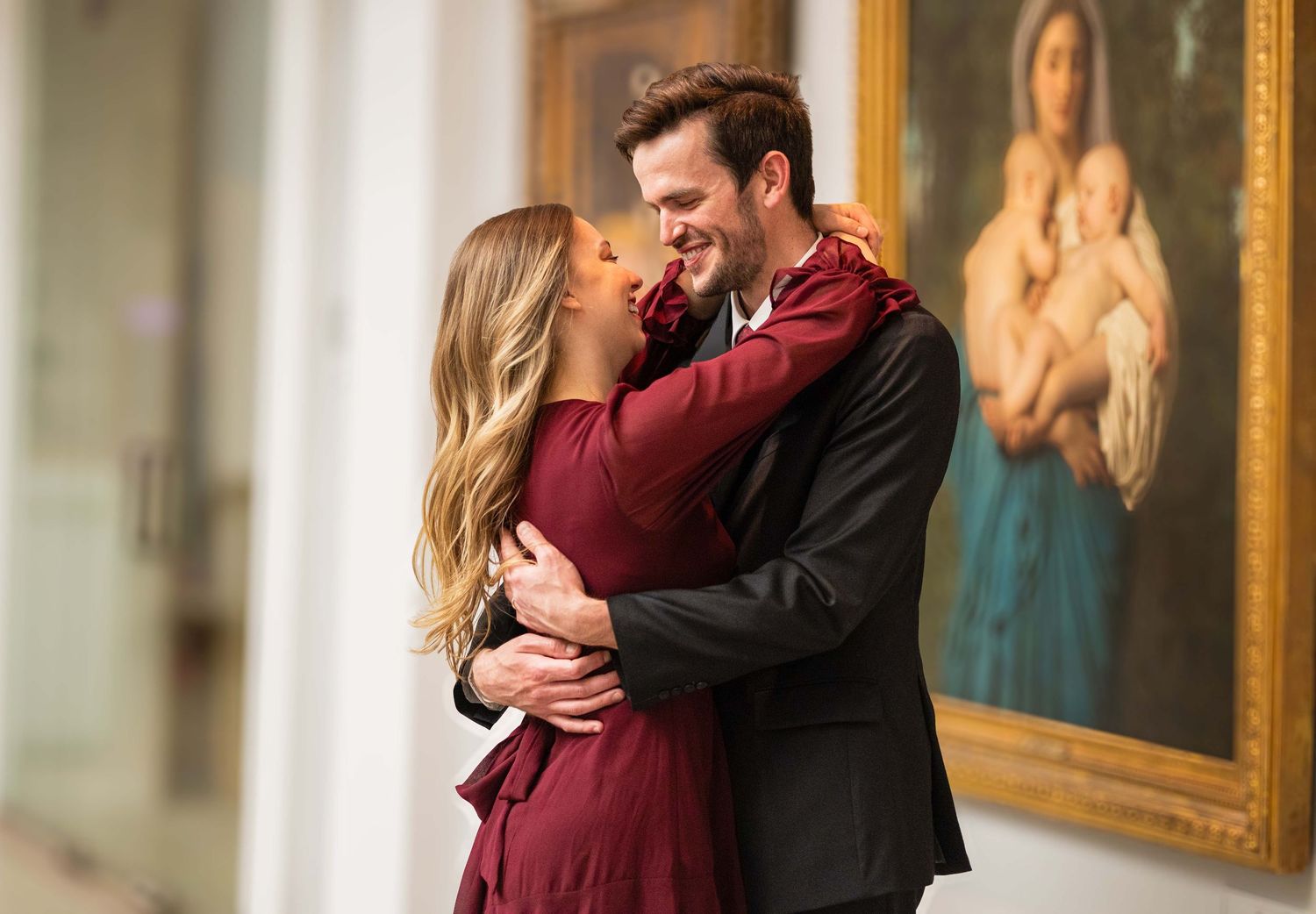 Romantic couple embracing and smiling in an elegant museum hallway with classical artwork backdrop.