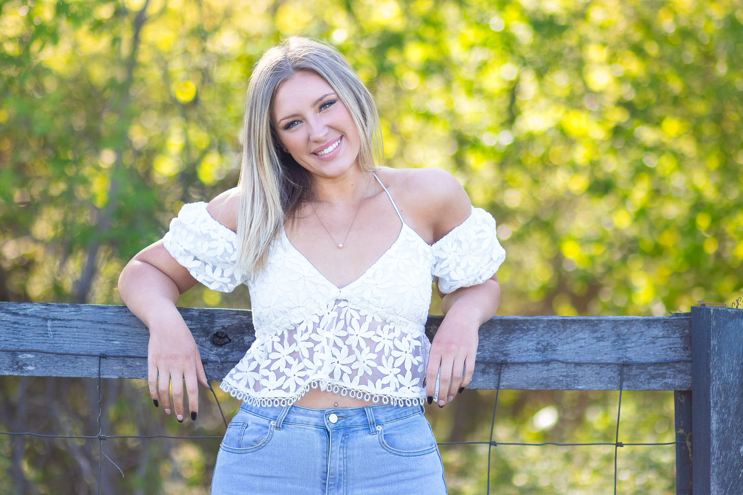 Smiling blonde woman in white lace crop top and jeans posing against a wooden fence with autumn foliage.