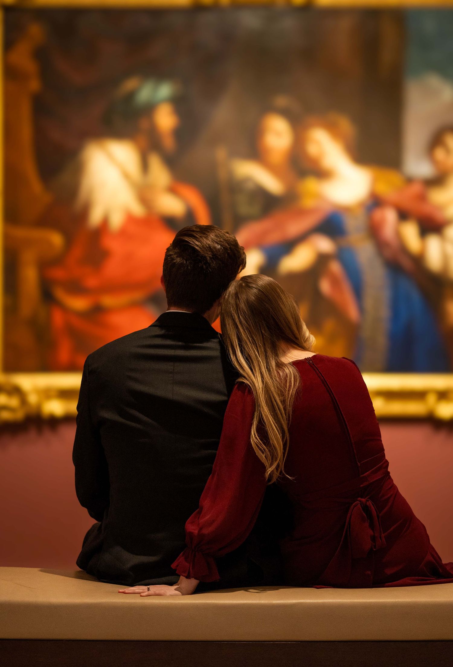 Couple sitting together on bench viewing classic painting in a museum gallery.