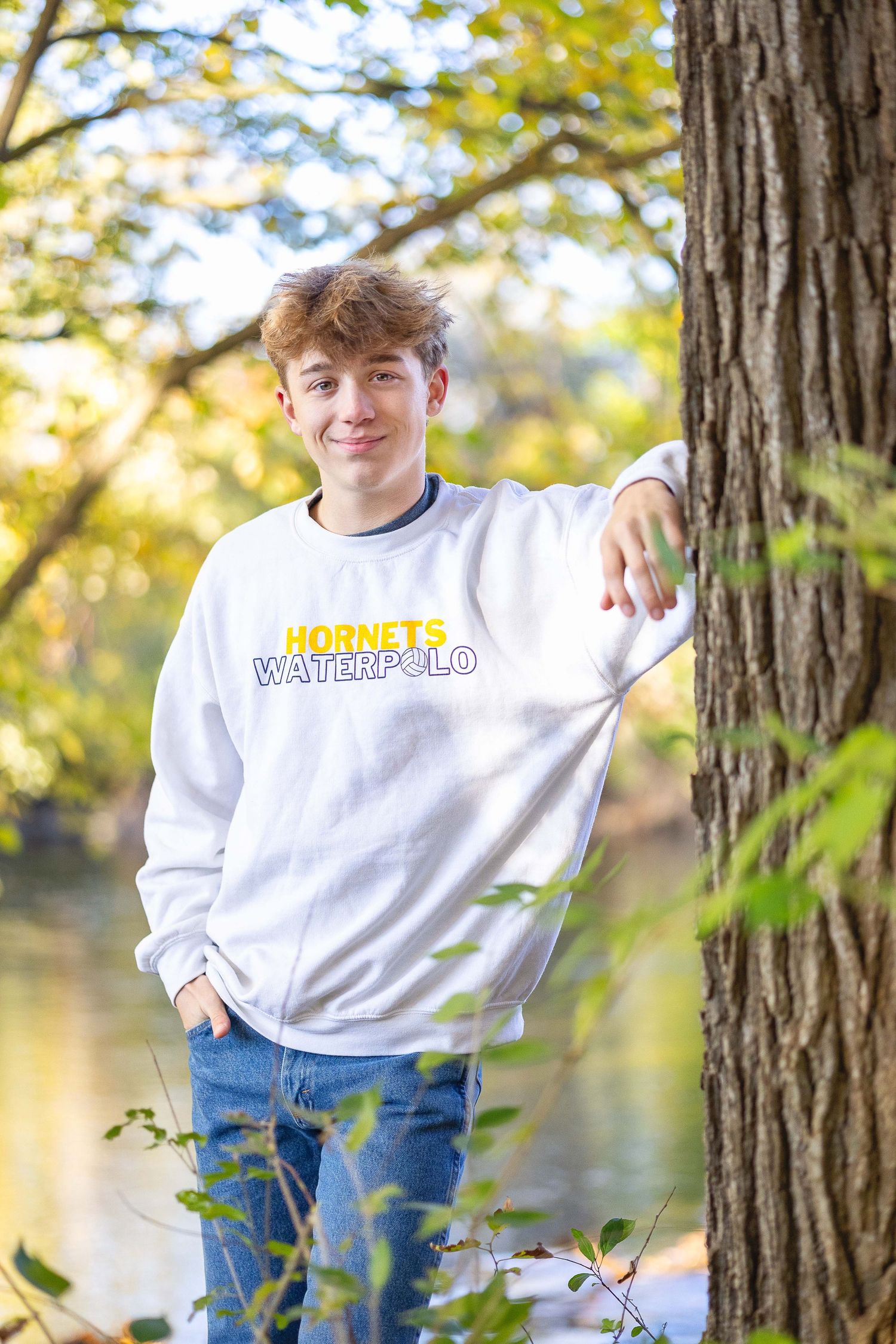 Teen boy in Hornets Water Polo sweatshirt leaning against tree near river in autumn park.