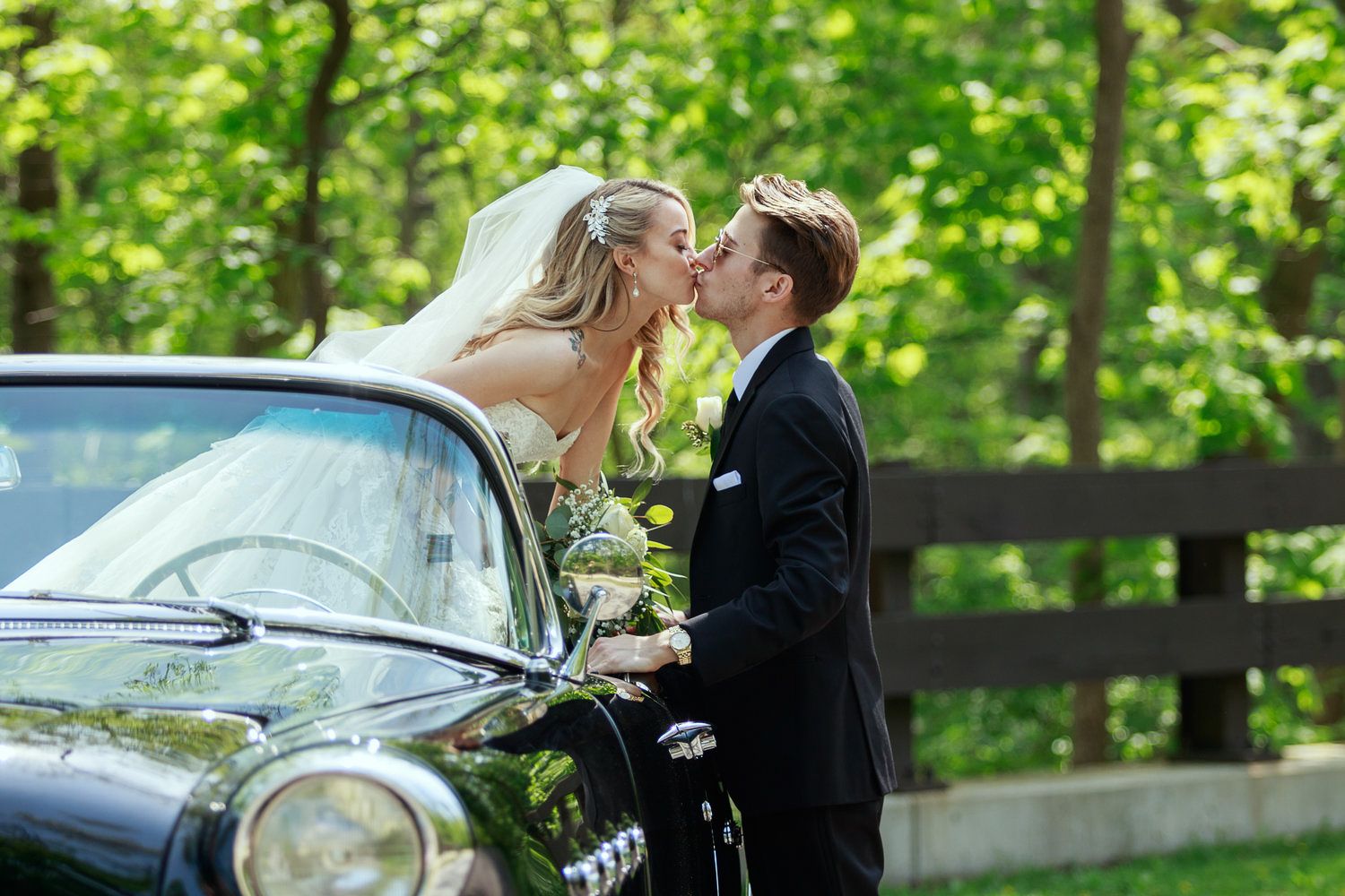 Bride in white gown and veil shares a romantic kiss with groom in black suit beside a vintage black car outdoors.