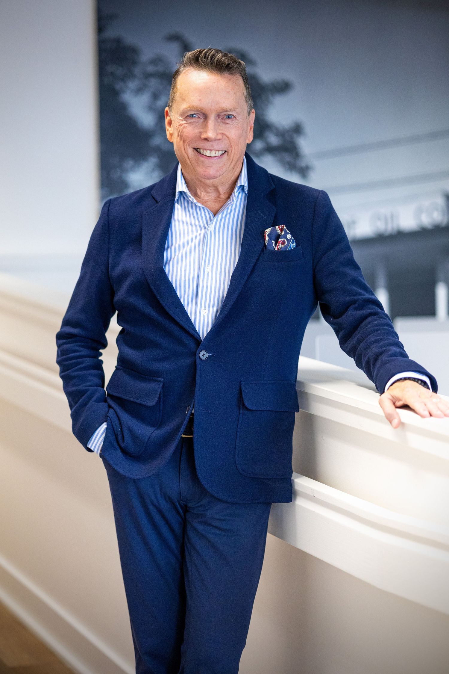 Smiling man in navy blue suit with pocket square leaning on white railing in modern interior setting.