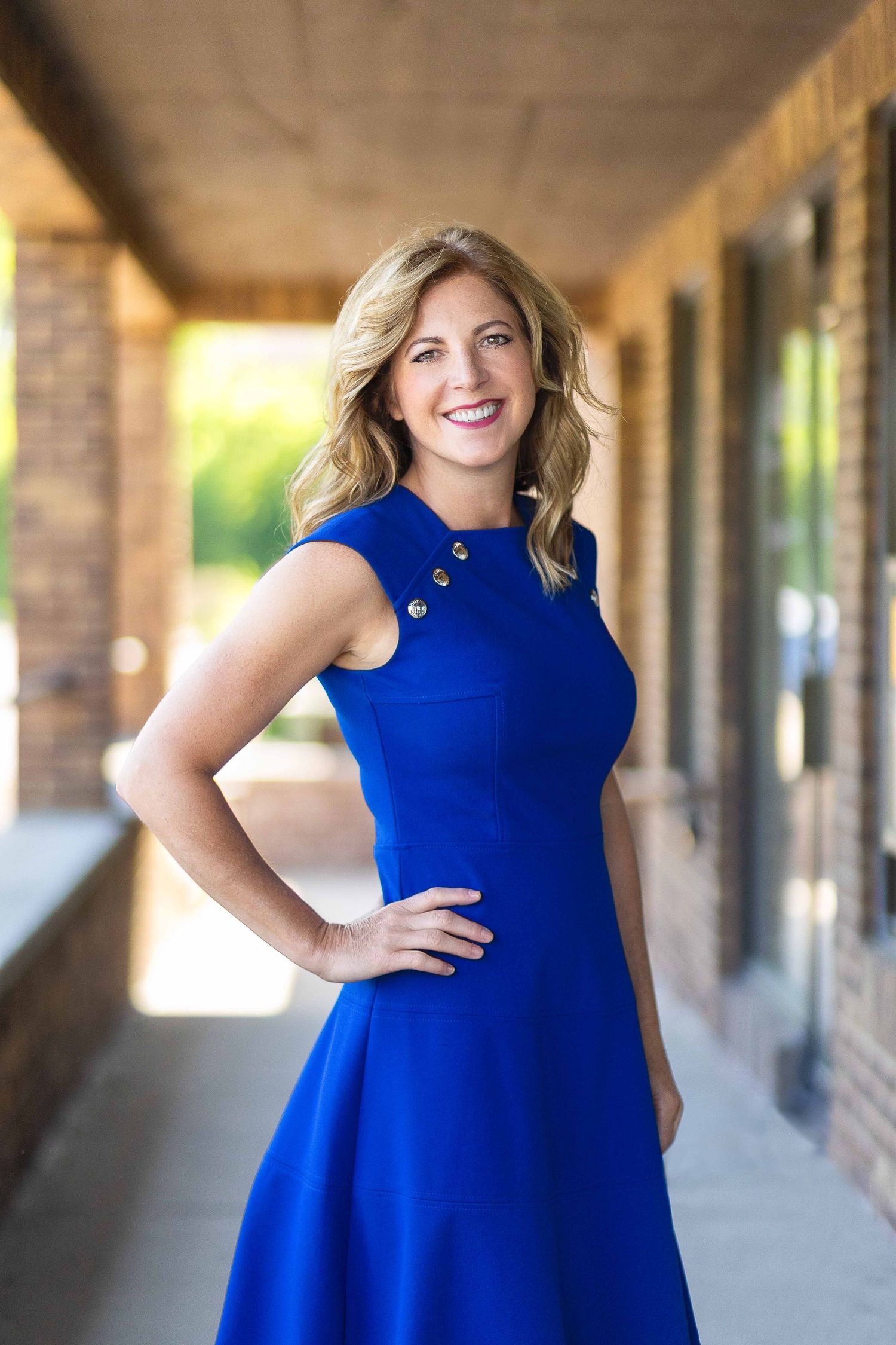 Confident blonde woman in royal blue dress posing for professional portrait along brick building corridor.
