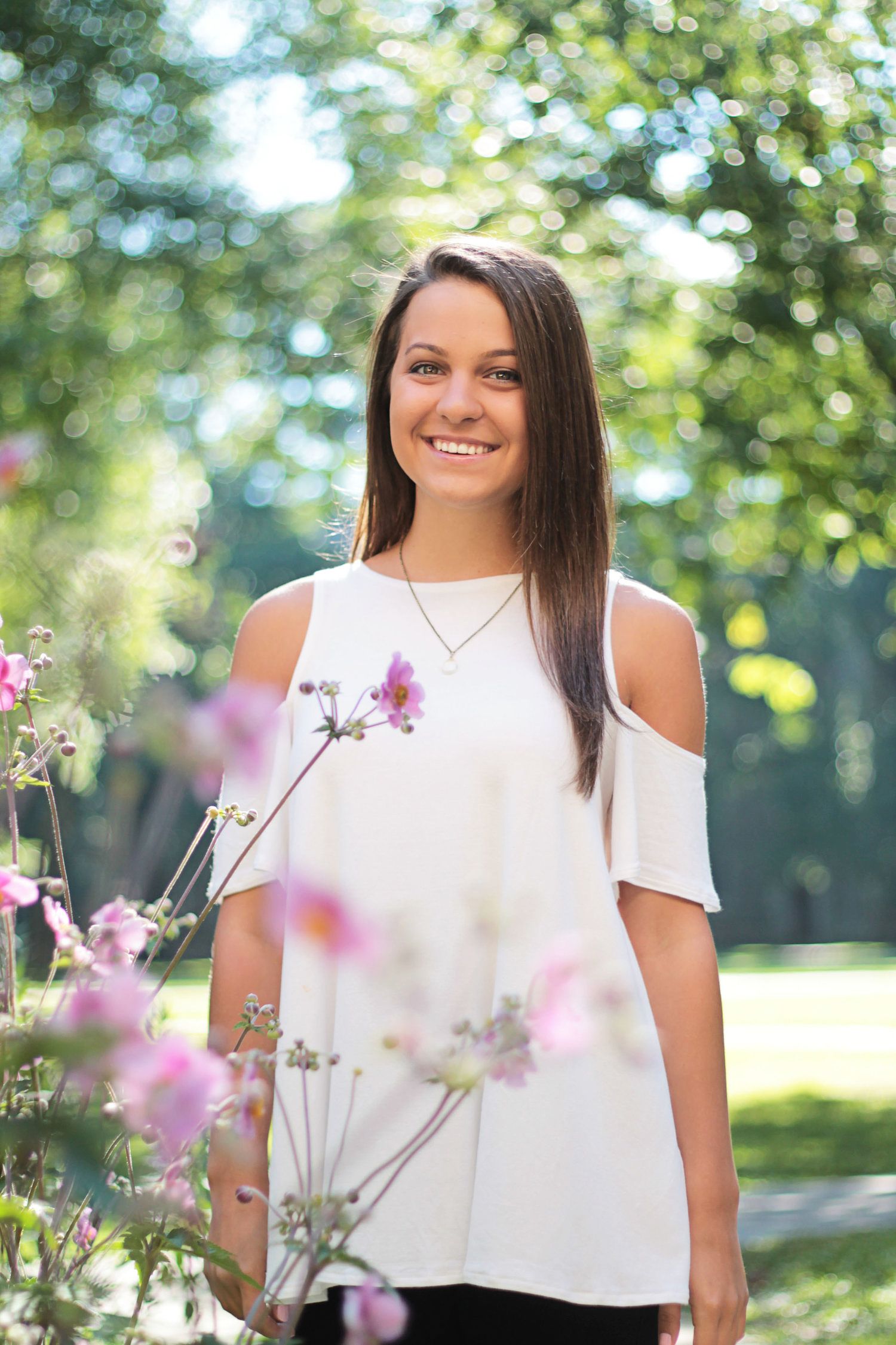 Smiling young woman in white cold-shoulder top posing near pink flowers in sunny green park.