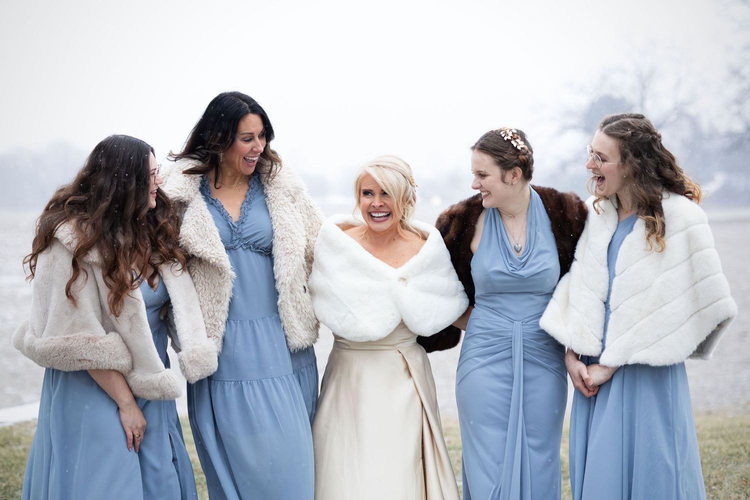A laughing bride and four bridesmaids in blue dresses with fur wraps enjoy a winter wedding.