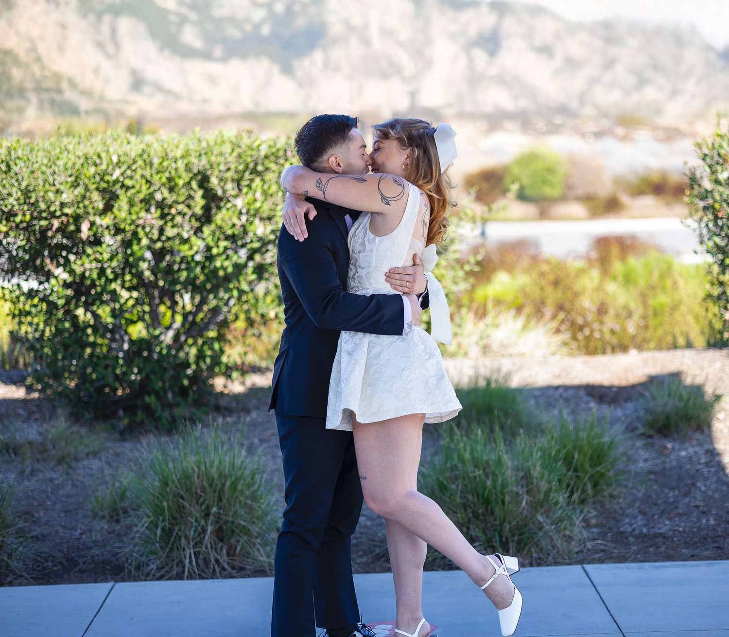 A newlywed couple shares a kiss outdoors, the bride in a short white dress and groom in a dark suit with mountains behind.