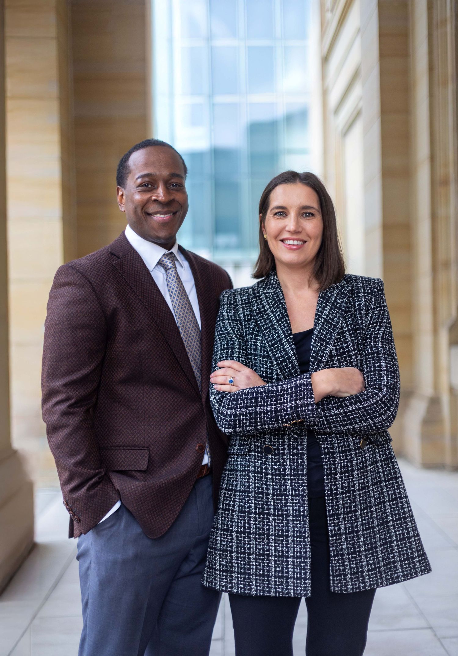 Professional man and woman in business attire smiling confidently in modern building corridor.