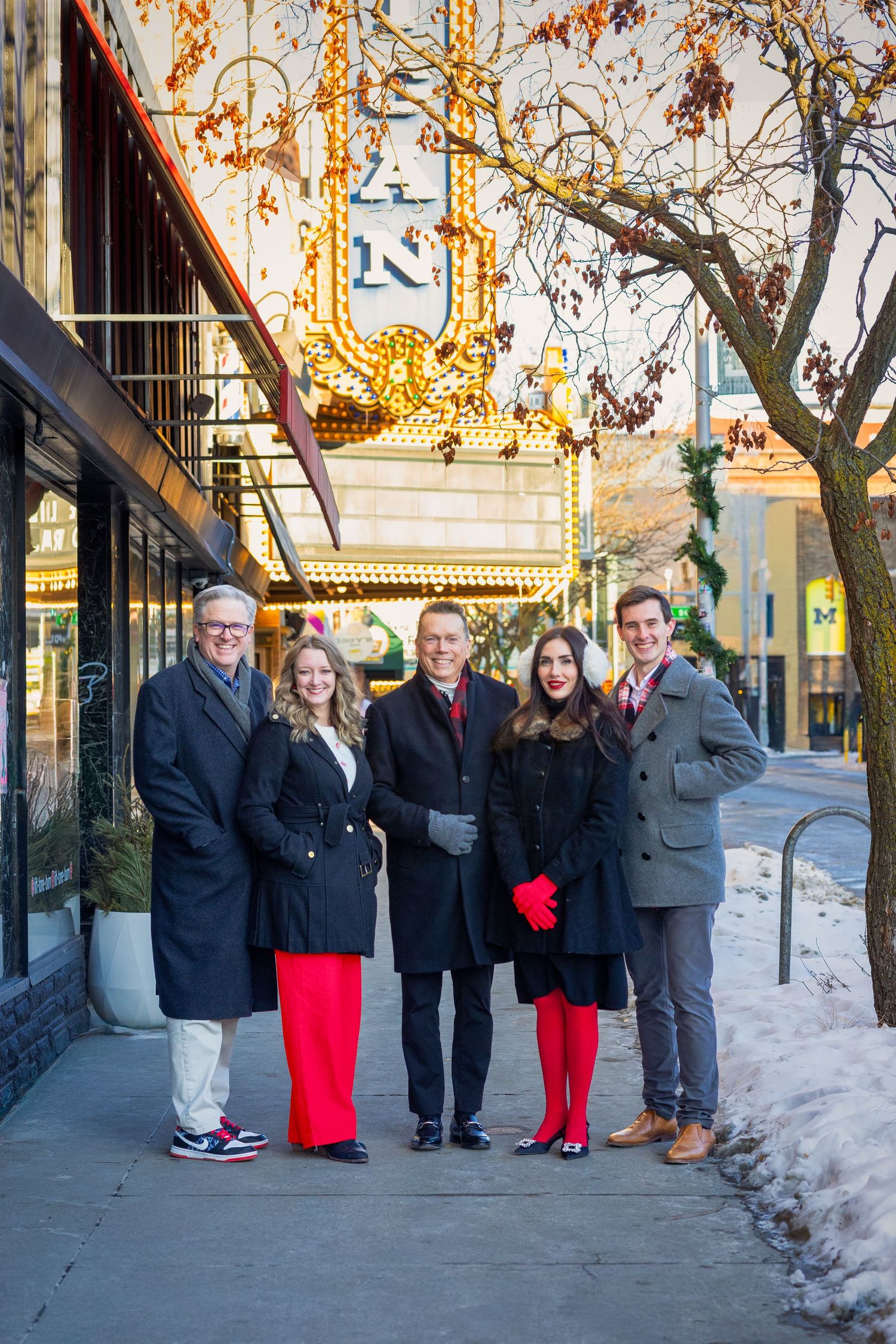 Group of five people in winter coats posing on snowy city sidewalk.