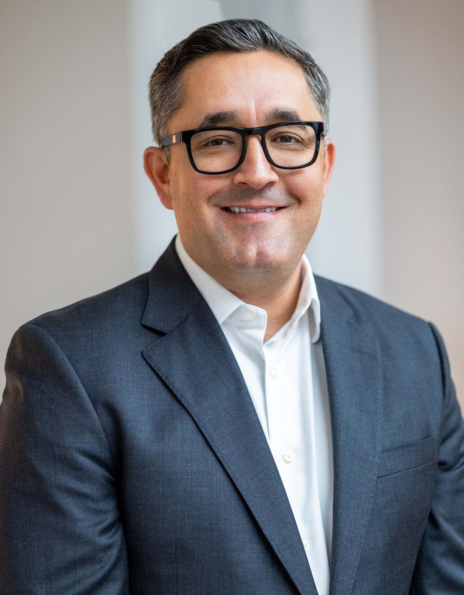 Professional middle-aged man in gray blazer and black glasses smiling for a corporate business headshot.