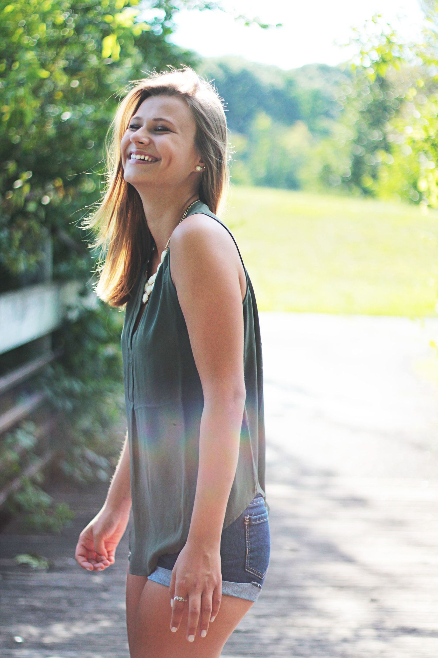 Young woman laughing outdoors in green sleeveless top and denim shorts on a sunny day.