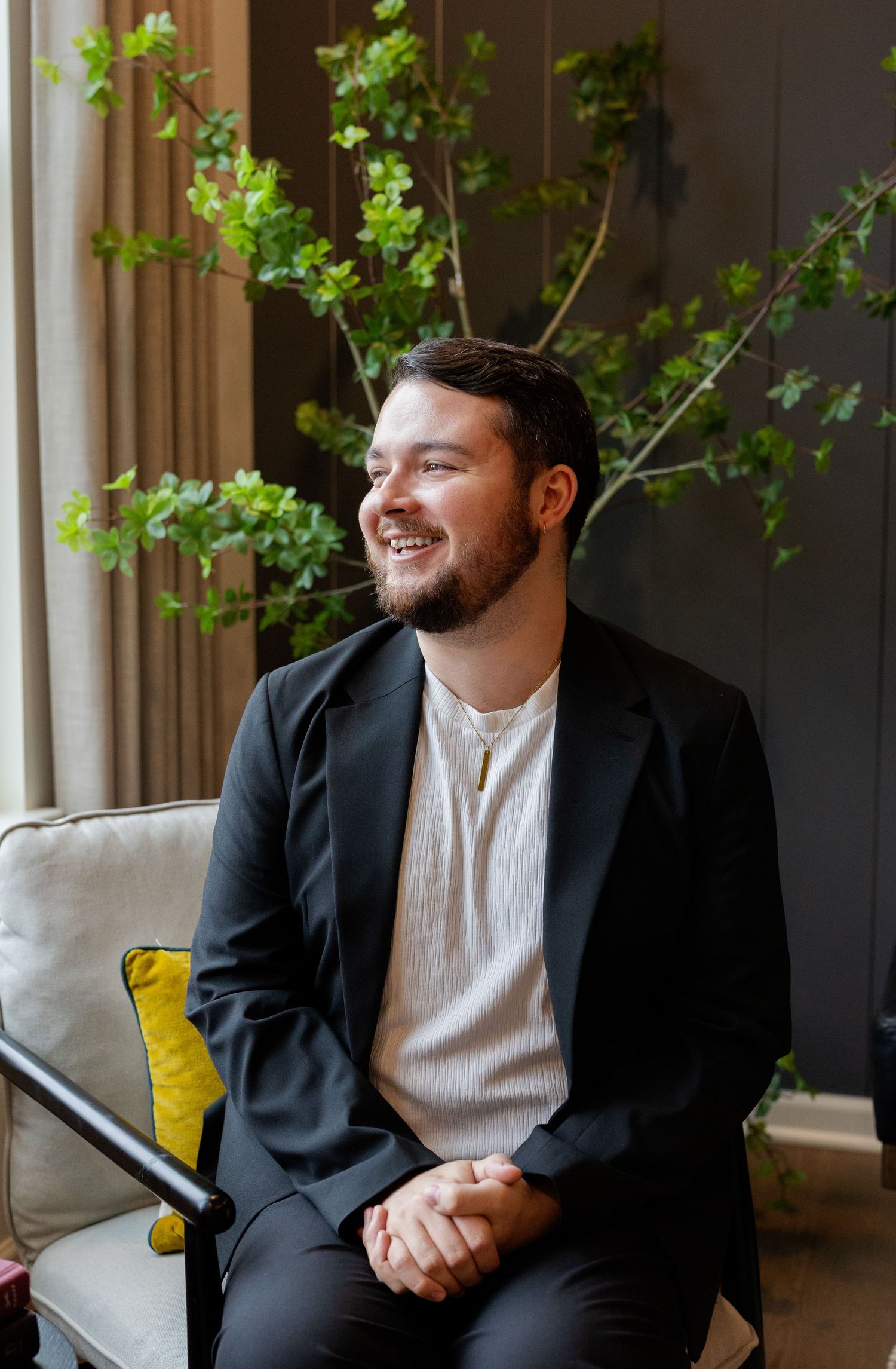 Young man in black suit smiling while seated near green plants indoors.