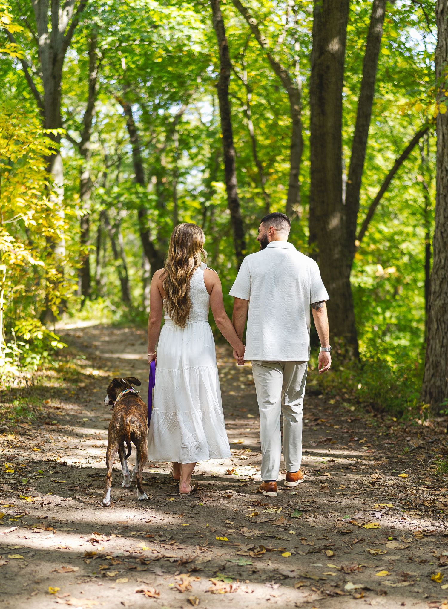 Couple holding hands walking dog on a sunlit wooded trail, woman in white dress and man in white shirt.
