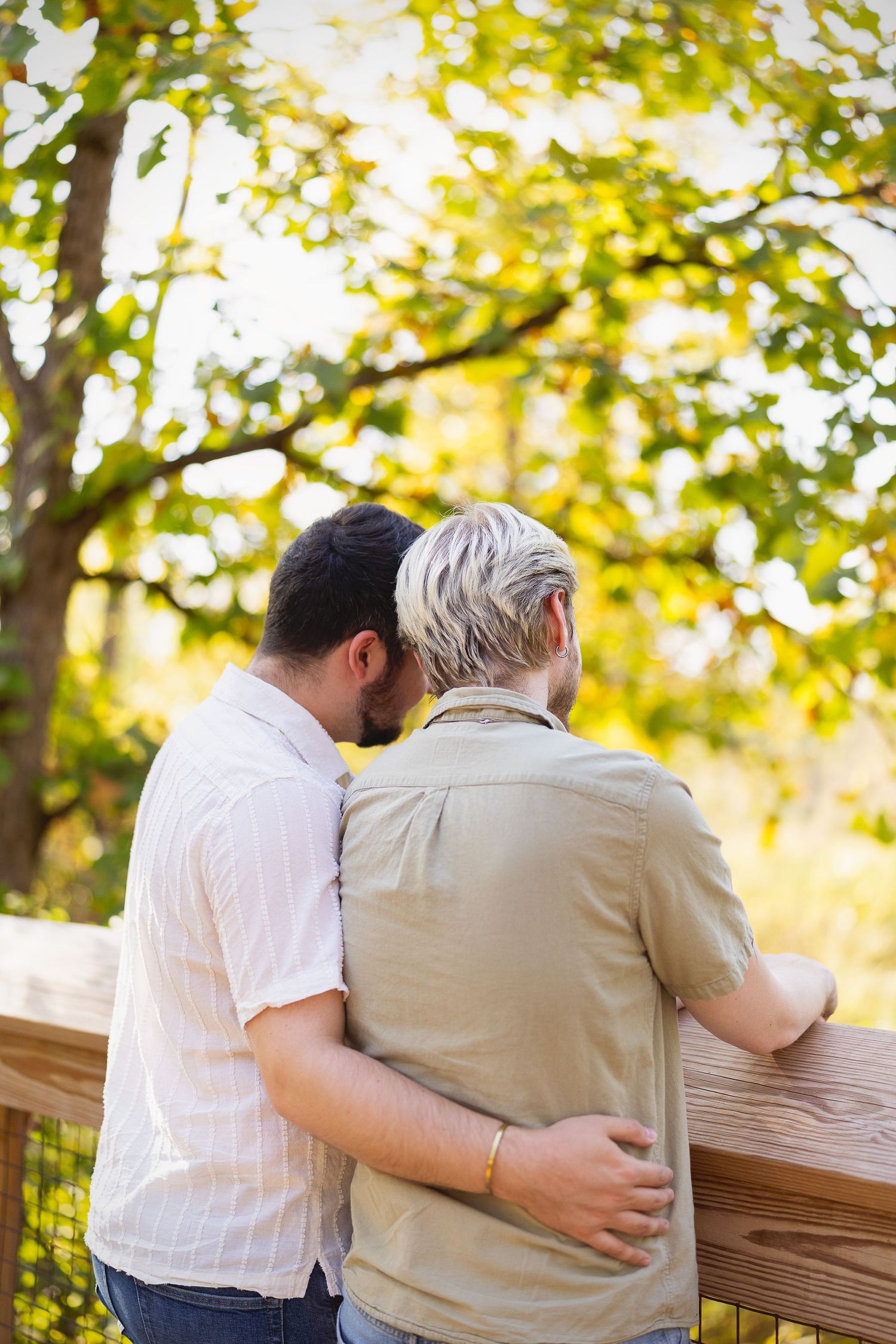 Couple embracing on wooden deck surrounded by lush green trees in warm sunlight.