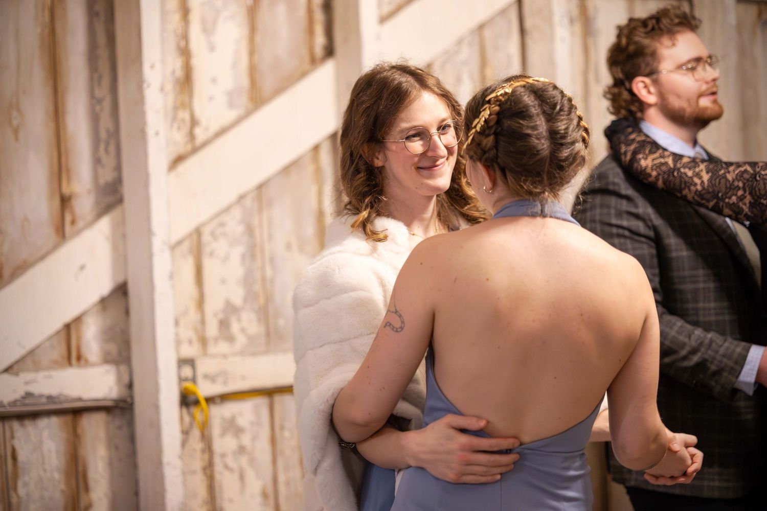 Bride in white wrap embracing bridesmaid in blue dress inside rustic barn venue.