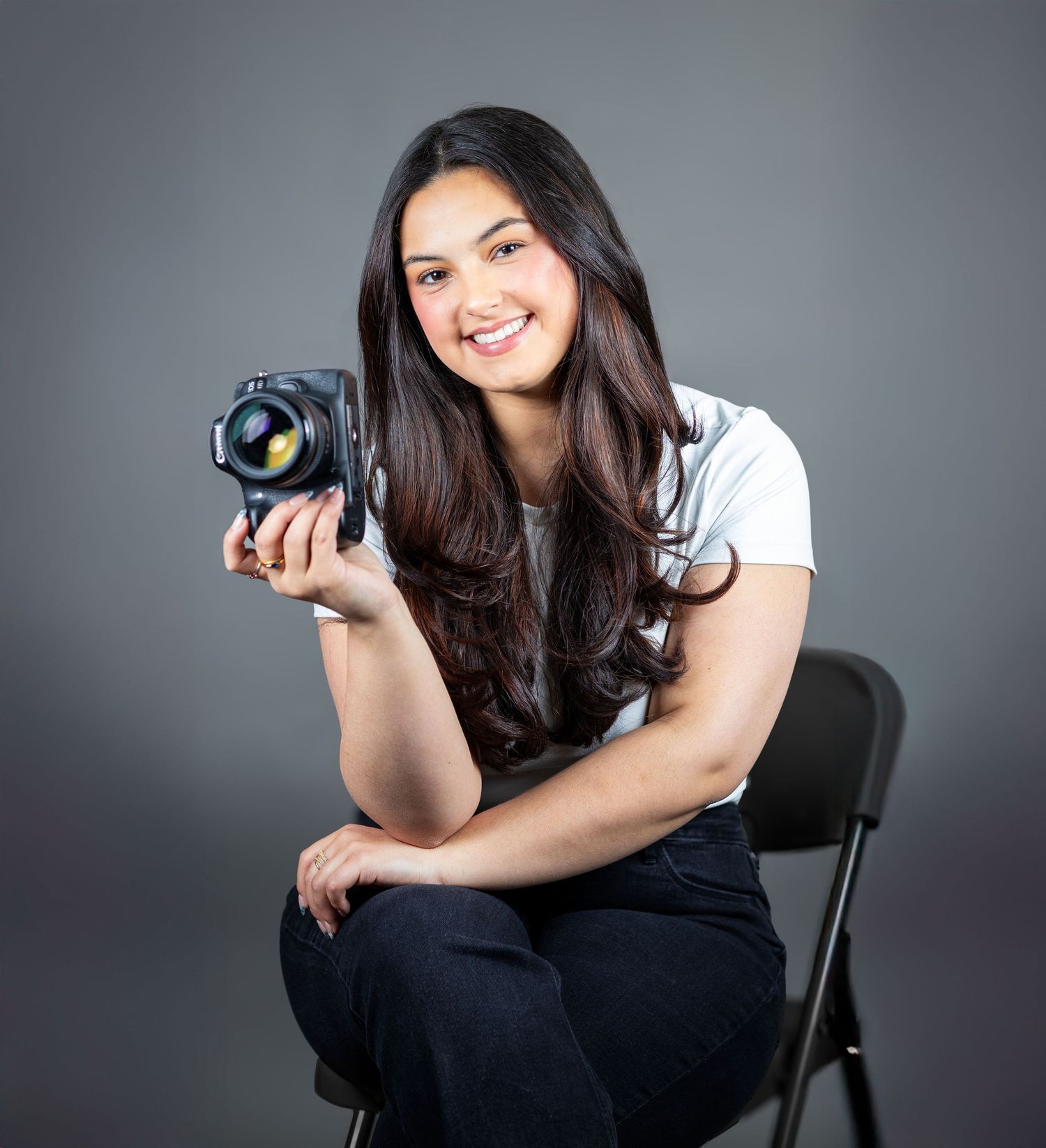 Young woman with long dark hair sitting on a chair holding a professional camera in a studio.