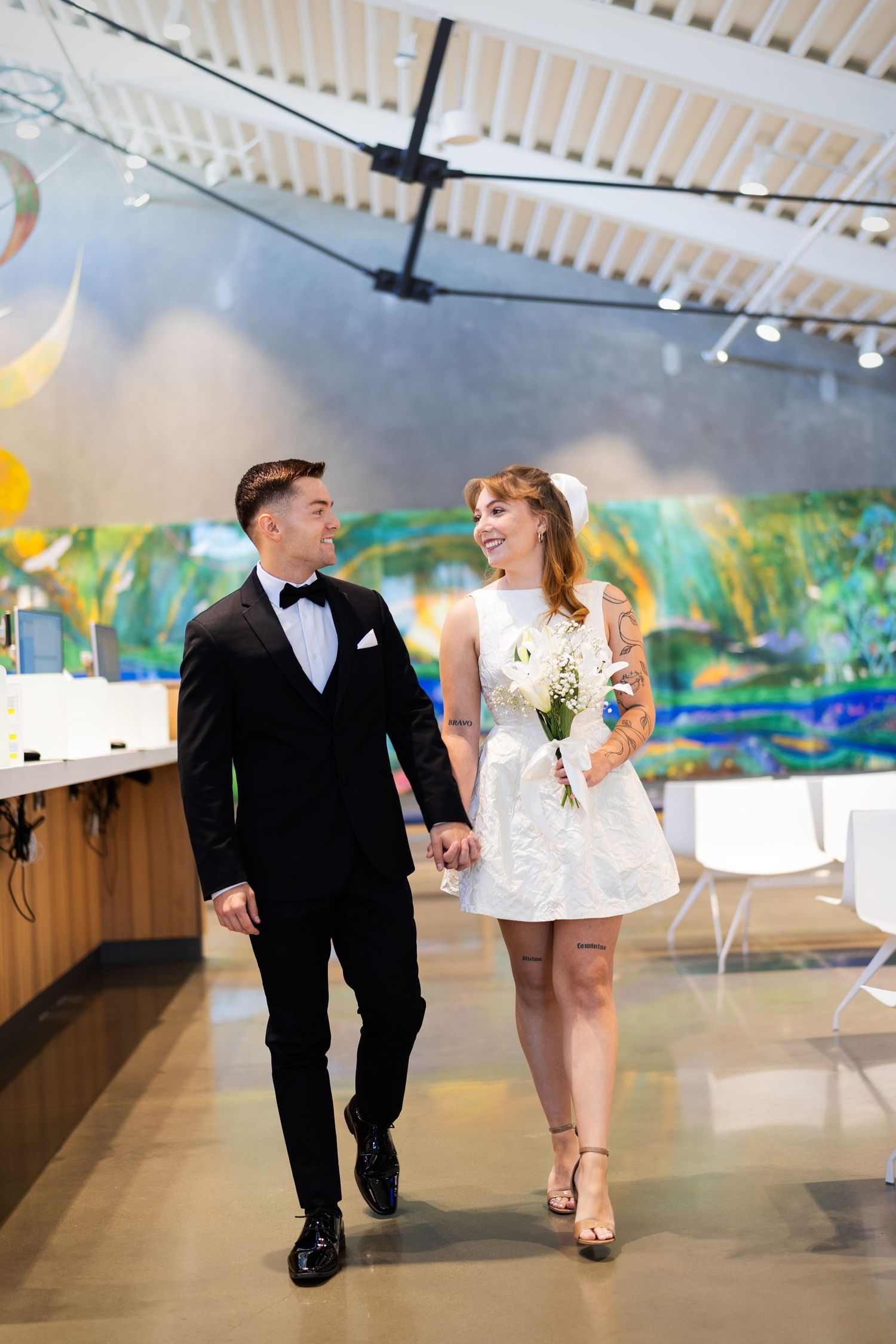Bride and groom in formal attire walking hand in hand inside a bright venue.