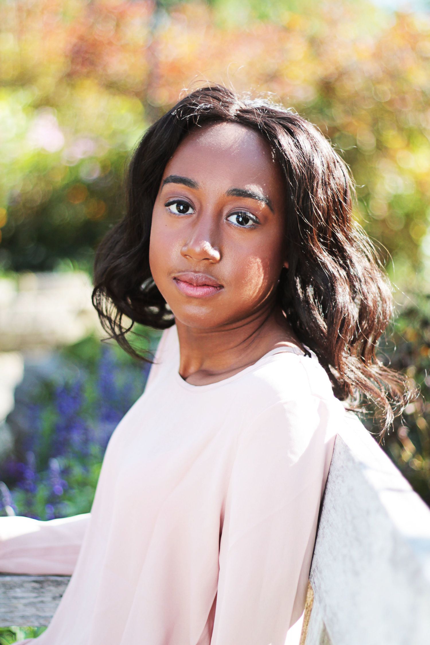 Young Black woman with wavy hair in blush pink top posing in colorful outdoor garden setting.