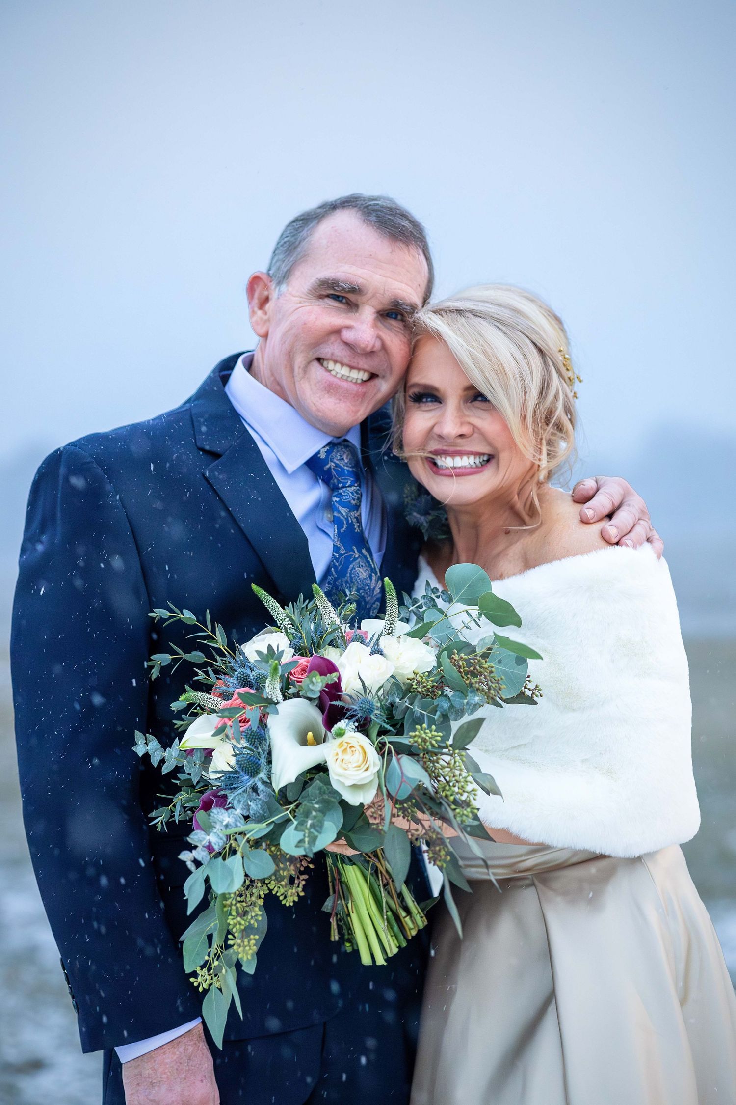 A smiling couple poses in the snow on their winter wedding day with a lush bouquet.