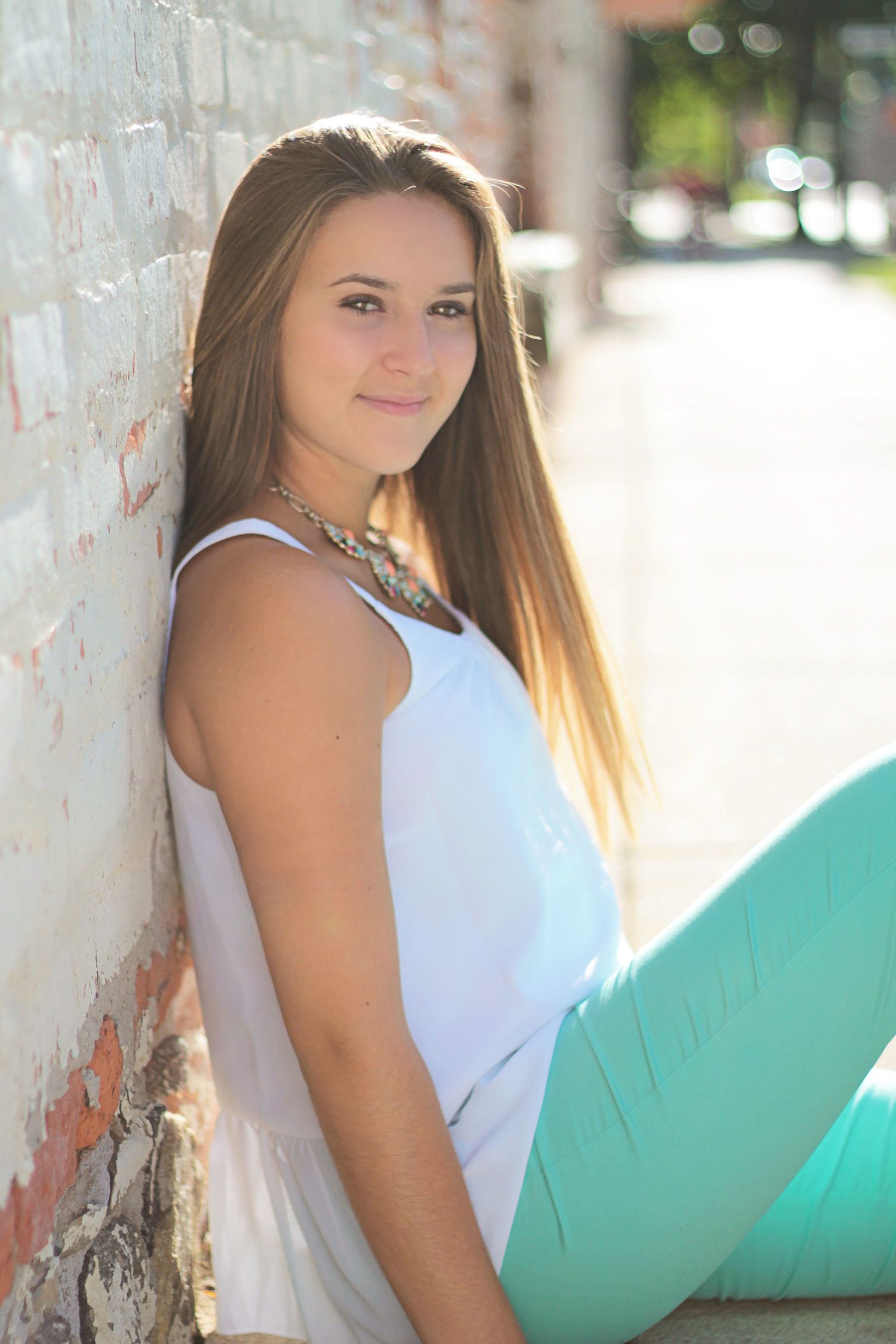 Young woman with long hair in white top and teal pants leaning against brick wall outdoors.