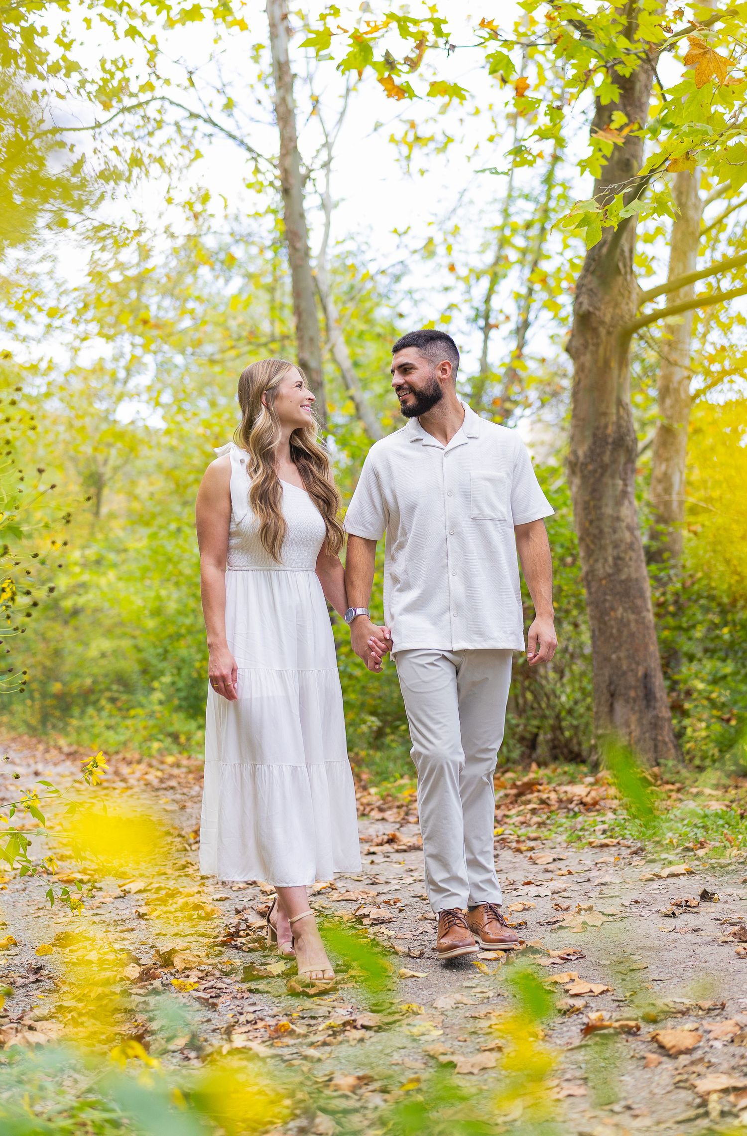 Couple holding hands walking on autumn leaf-covered path surrounded by colorful fall foliage trees.