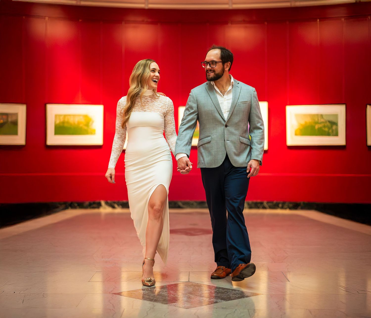 Elegant couple holding hands and smiling while walking through a red-walled art gallery.