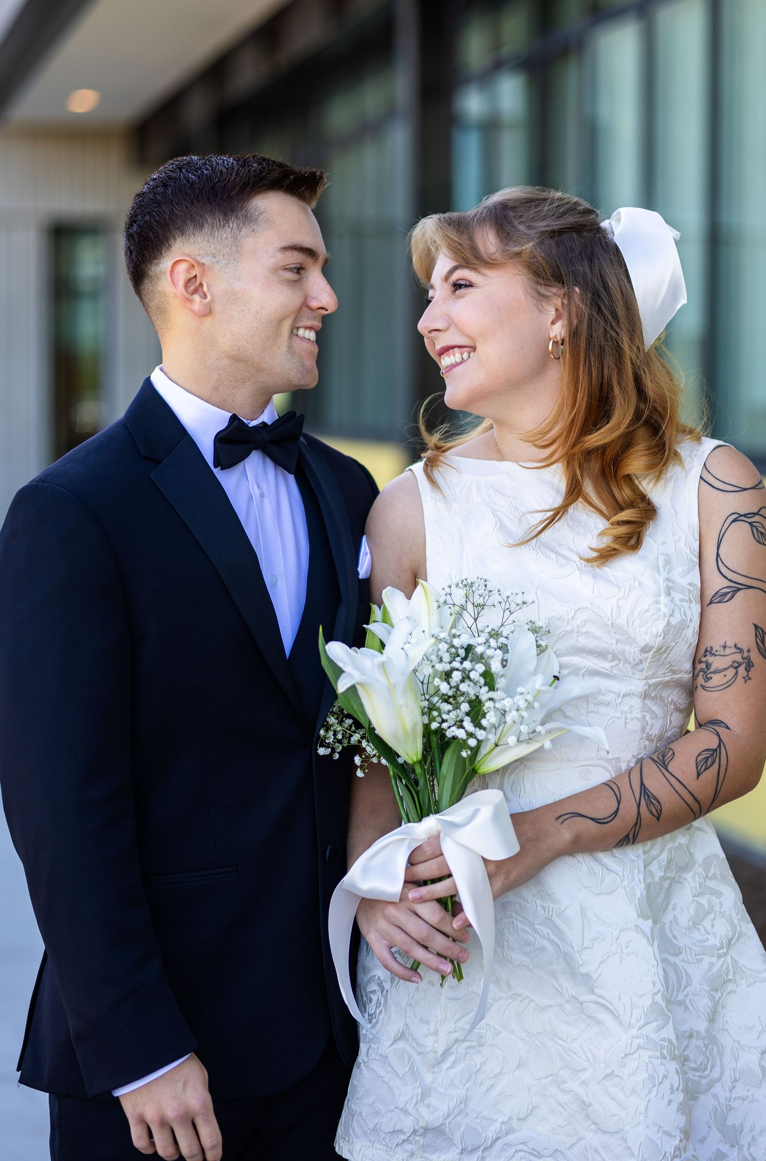 Tattooed bride holding white bouquet smiling at groom in black tuxedo outdoors.
