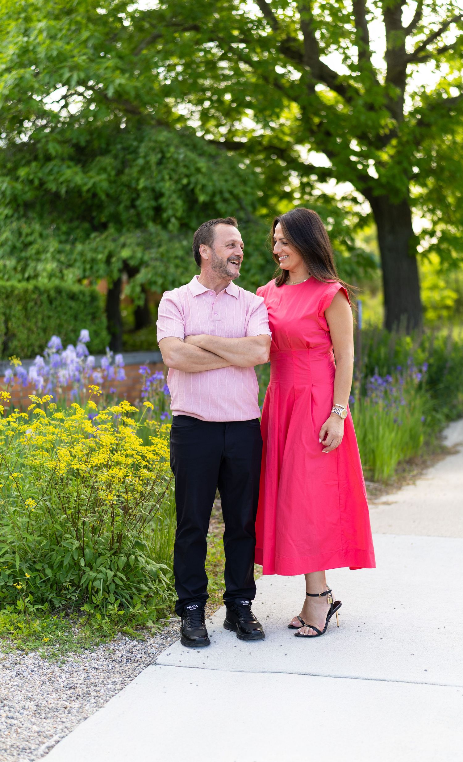 Laughing couple standing together on garden path surrounded by colorful blooming flowers.