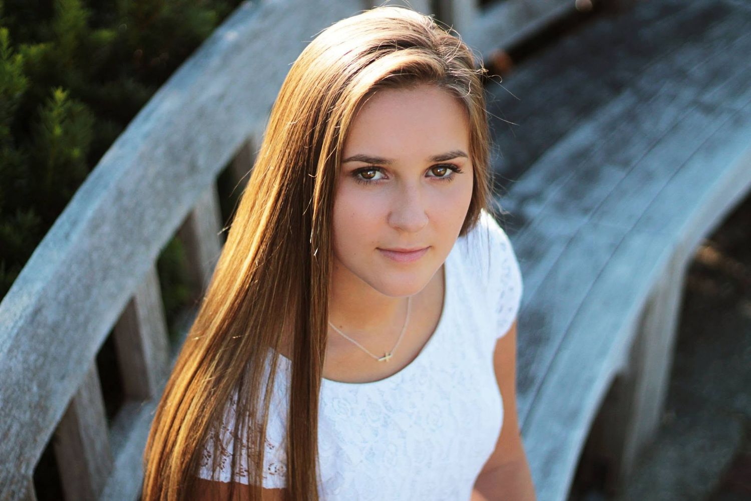 Young brunette woman with long hair in white lace top posing on weathered wooden bench.