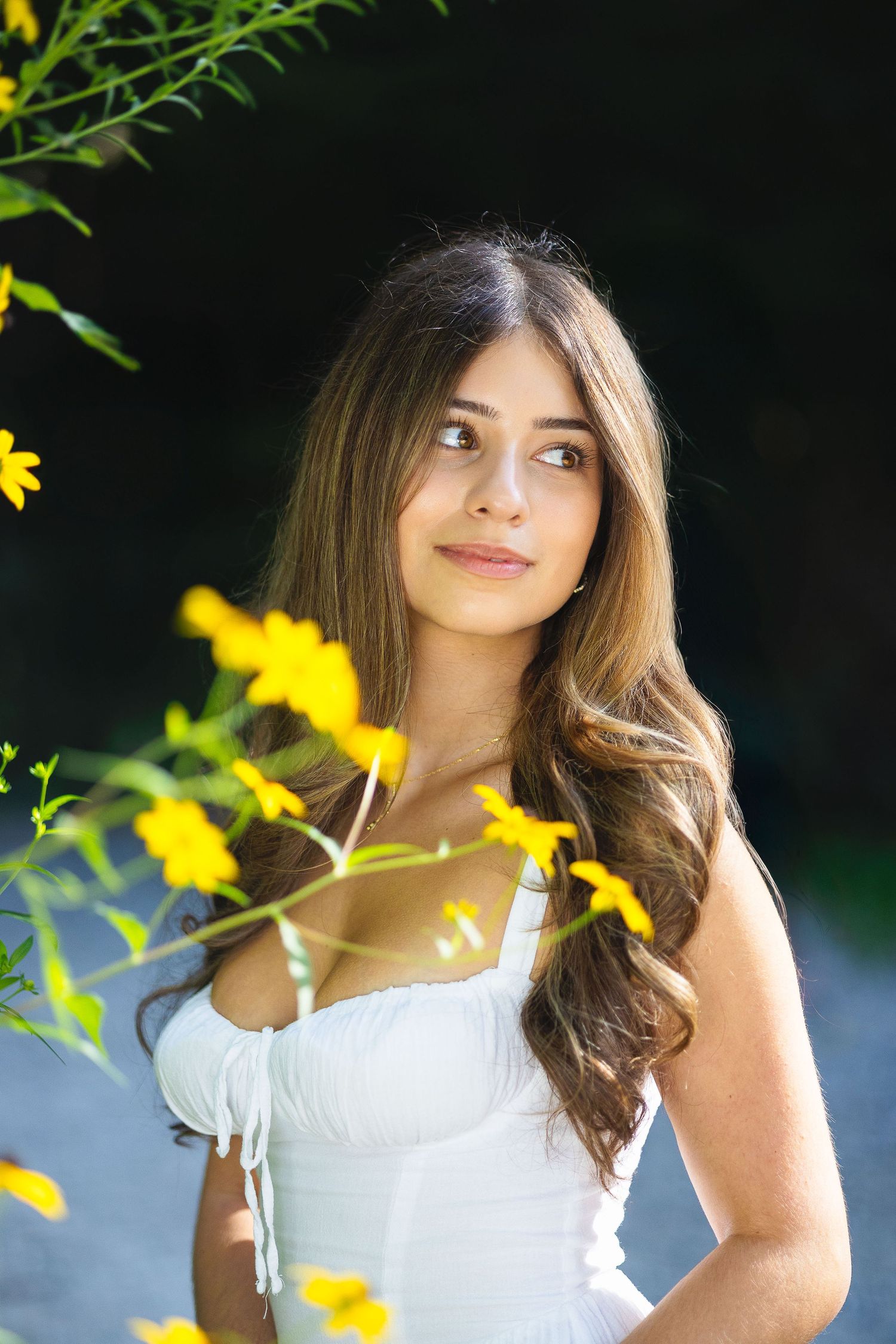 Young woman with long brown hair in white dress poses near yellow wildflowers in sunlit garden.
