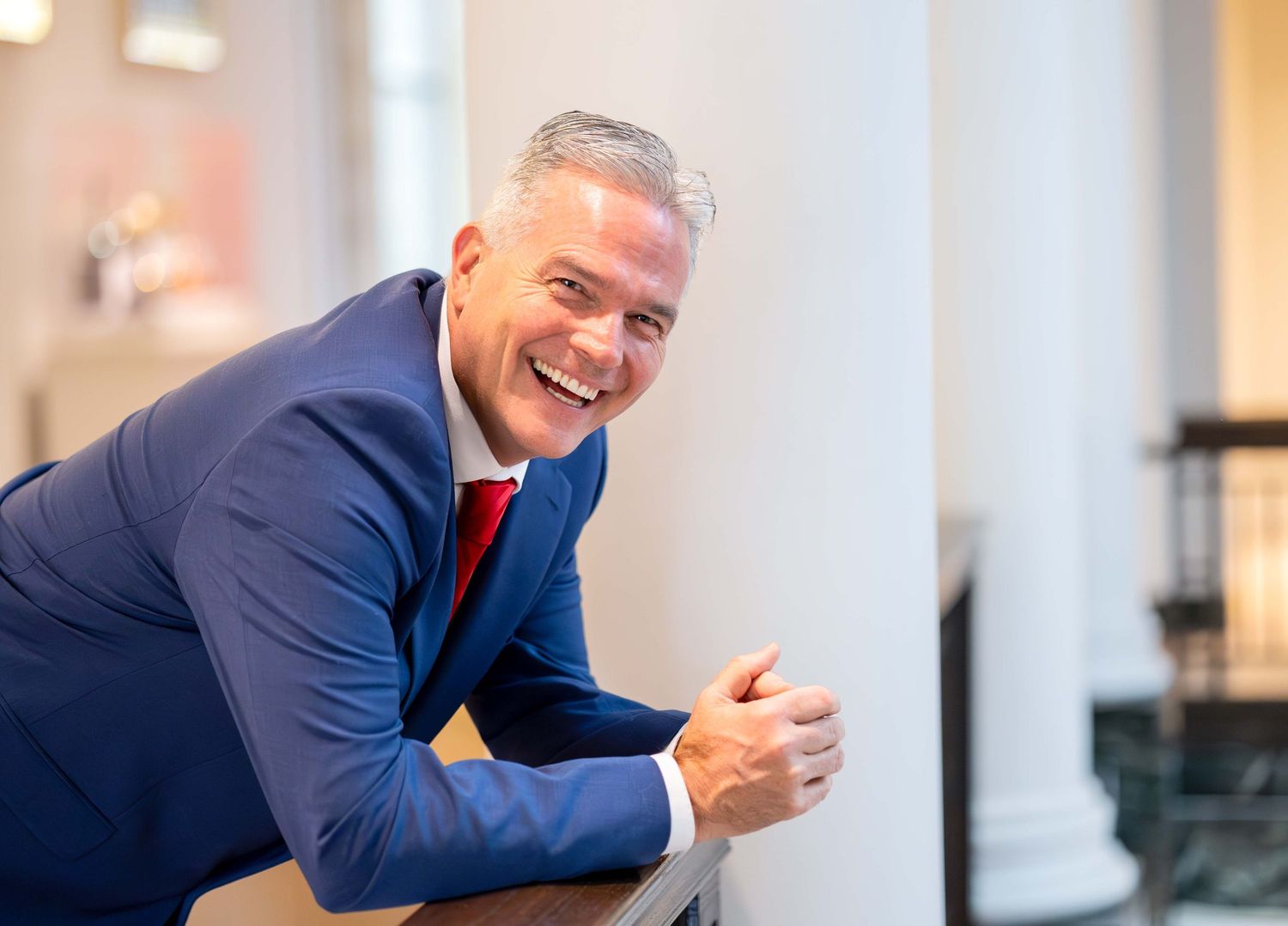 Cheerful professional man in blue suit and red tie leaning on white column for business portrait.