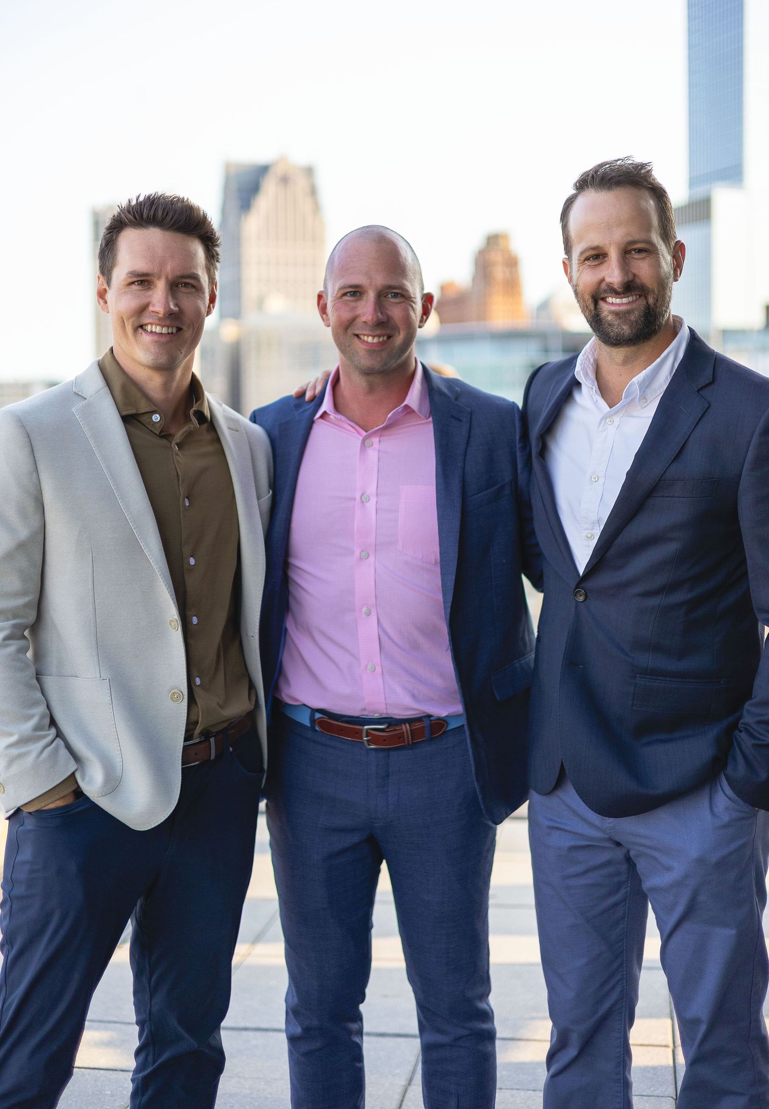 Three smiling men in blazers posing together on a rooftop with a city skyline at sunset.