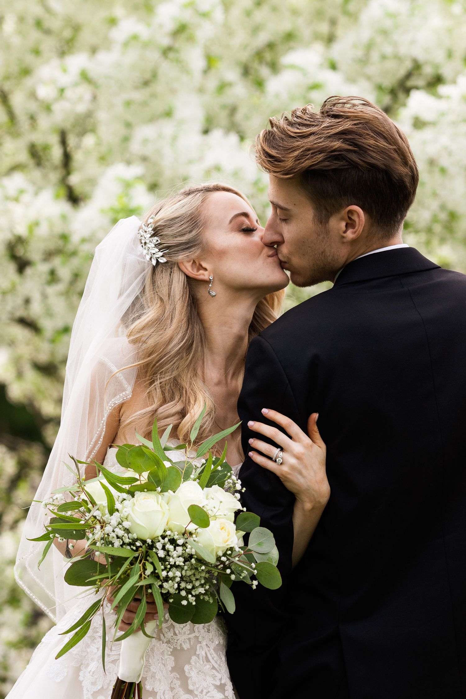 Bride in lace gown and veil kisses groom in black suit while holding a white rose and greenery bouquet outdoors.