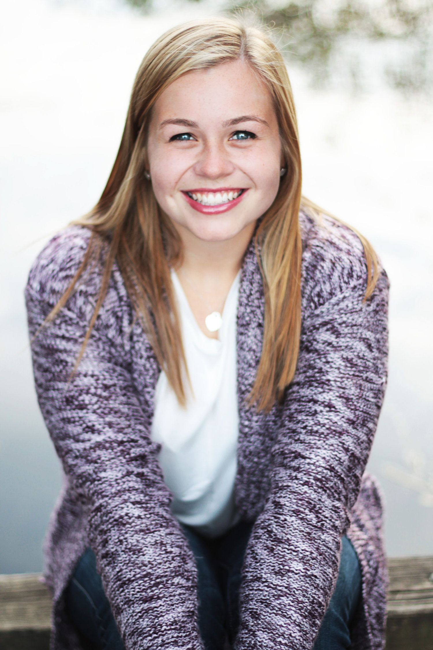 Happy teenage girl with blonde hair in purple cardigan sitting outdoors near water smiling.