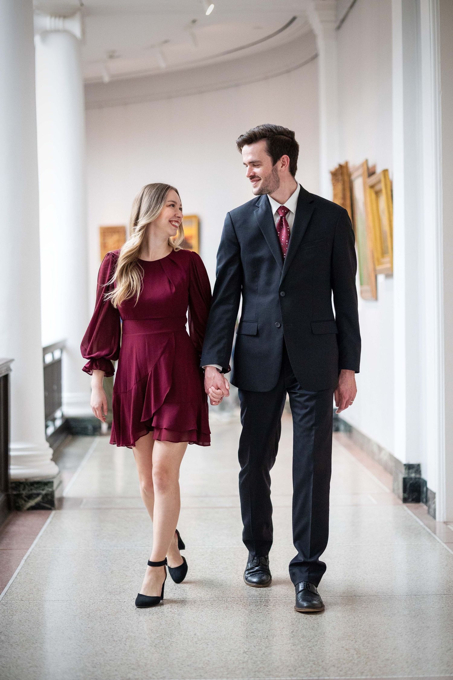 Couple walking hand in hand through elegant museum hallway, smiling at each other.