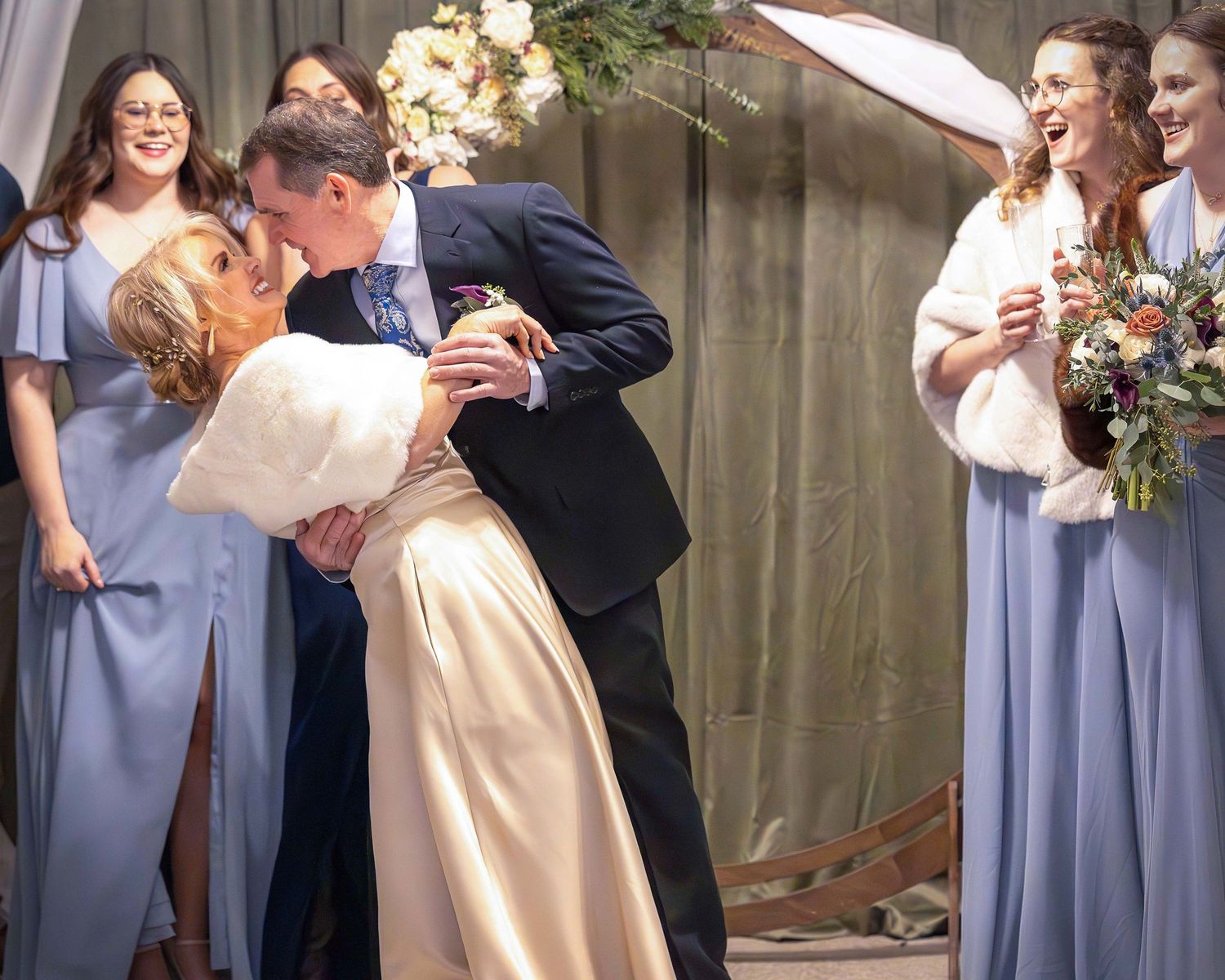 Bride and groom share a romantic dip kiss surrounded by laughing bridesmaids in blue dresses.