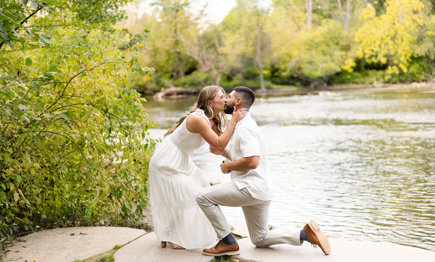 A couple shares a tender kiss during a marriage proposal by a serene riverside.