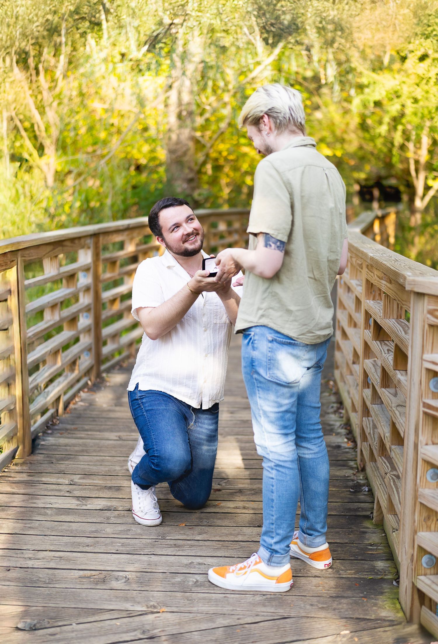 A man kneels on a wooden bridge proposing to his partner in a sunlit outdoor setting.